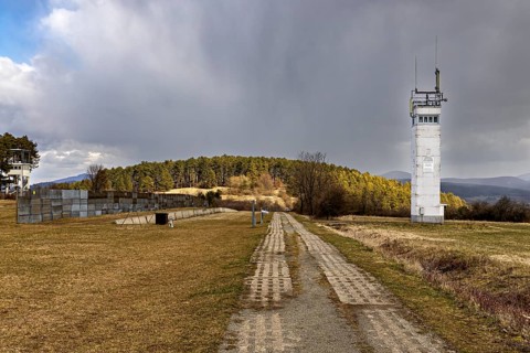 A white watchtower stands next to a long path in a meadow with a cloudy sky, the Point Alpha