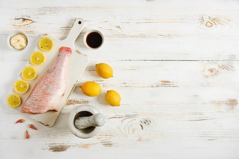 Raw red snapper is placed on cutting board next to sliced lemons and bowl of spices. On side are