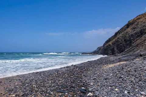 Pebble beach at the base of high cliffs, ocean waves rolling in, the sky is cloudless blue, Rogil,