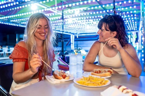 Two young women are laughing and eating hot dogs and french fries at a table, enjoying fast food