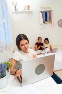 Teenager concentrating on her laptop at a desk, studying in her bedroom while her young sisters