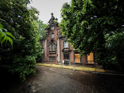 An old brick villa stands abandoned and boarded up with wooden panels behind a construction fence,