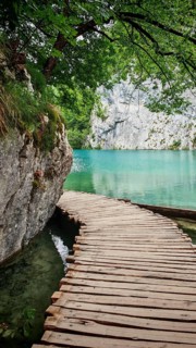 A wooden walkway winds along a turquoise lake, framed by rocks and trees, Plitvice Lakes National