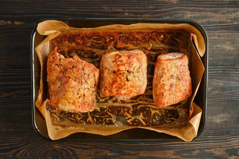 Pork fillets on baking sheet with parchment paper. Seasoning and herbs are visible on meat.