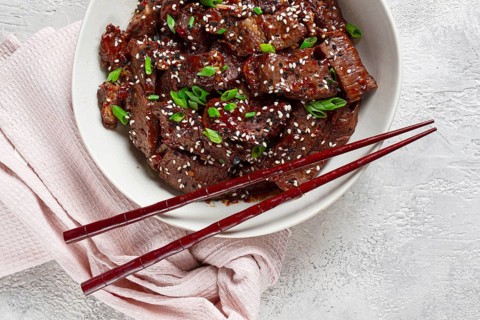 Crispy beef, with green onions and sesame seeds, in a bowl with chopsticks, on a textured surface,