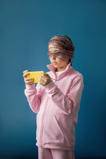 A child stands absorbed in a smartphone, eyes covered by tangled colorful wires, symbolizing the