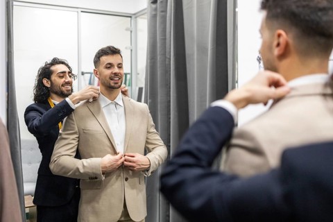 A tailor skillfully adjusts a client suit jacket in a fitting room. The client smiles while the