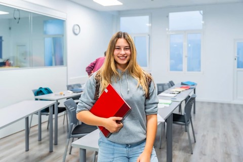 Young female student smiling while holding her red notebook and looking at the camera, standing in