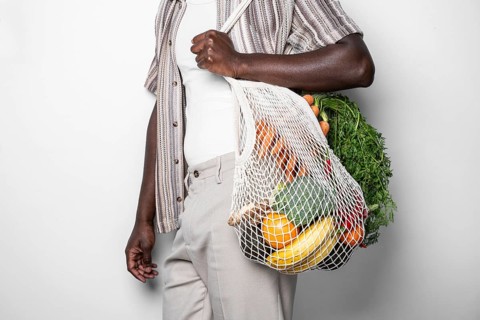 A close-up image of a man holding a reusable mesh bag filled with various fresh vegetables and