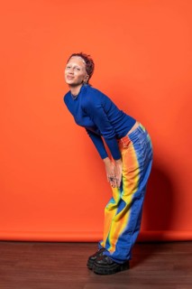 A fashionable woman poses against an orange backdrop wearing vibrant, multicolored trousers and a