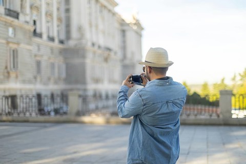 A Latino transgender man in a denim jacket and hat captures photographs of city architecture The