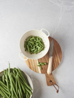 Fresh raw pod green beans cut into chunks in a colander next to a knife, the ends on a wooden
