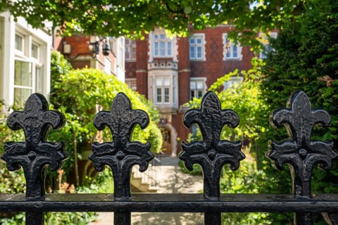 Detail, decorated wrought iron gate painted black in front of a garden and a brick building in the