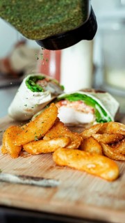A close-up of crispy fried potato wedges garnished with herbs alongside a fresh chicken wrap, set