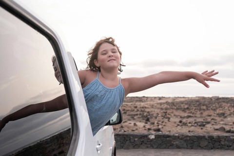 A young girl joyfully leans out of a car window, feeling the breeze as she embraces the spirit of a