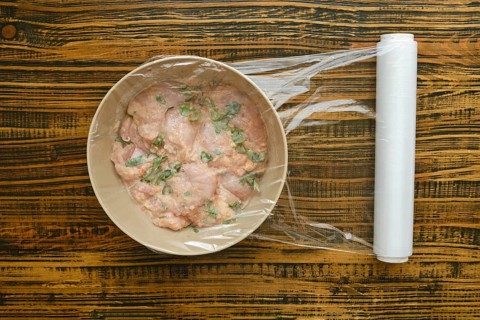 Chicken pieces mixed with herbs, garlik, paprika in bowl on wooden table. Clear wrap covers the