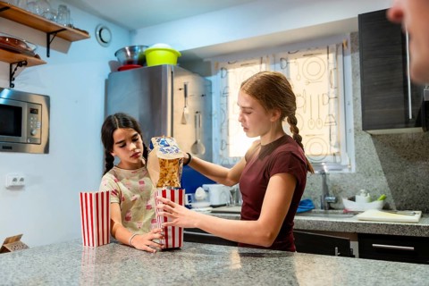 Two young sisters standing in a kitchen, one pouring popcorn from a bag into striped paper
