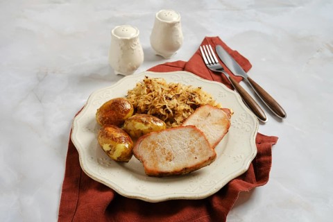 Plate with sliced roasted pork chops, roasted baby potatoes, and fried sauerkraut on table. Set