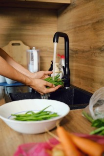 Hands hold green beans under running water in kitchen sink before meal preparation. Bowl with more