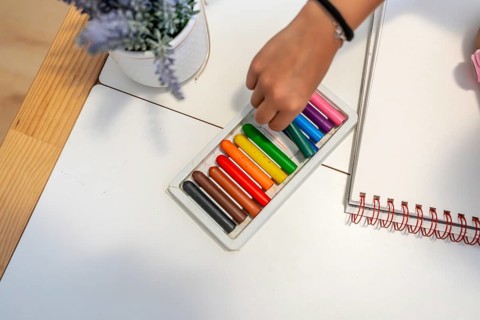 Child's hand selecting wax crayons from a box, preparing to draw on a blank notebook, symbolizing
