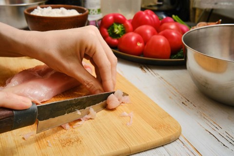 Close-up view of cutting chicken breast on a wooden board in a kitchen. Fresh tomatoes and a bowl