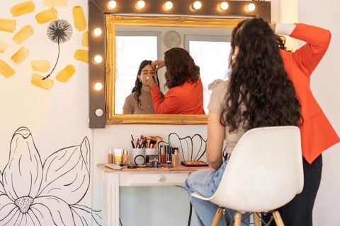 Rear view of a makeup artist working with a seated client, both reflected in a Hollywood-style