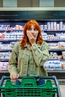 Woman standing in a supermarket aisle with a shopping cart, looking surprised and worried about the