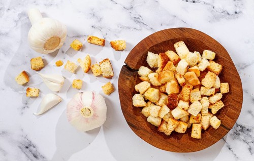 Bread is cut into small pieces and toasted until golden. Garlic sits nearby on a kitchen counter