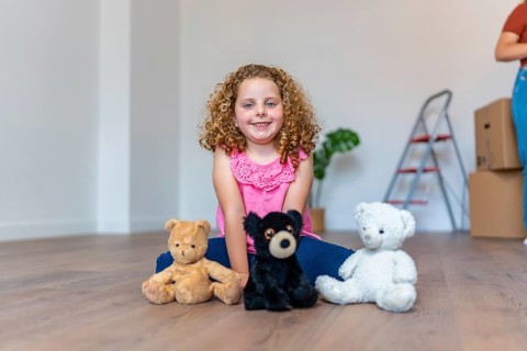 Happy curly haired girl sitting on the floor hugging teddy bears in an empty new home, smiling with