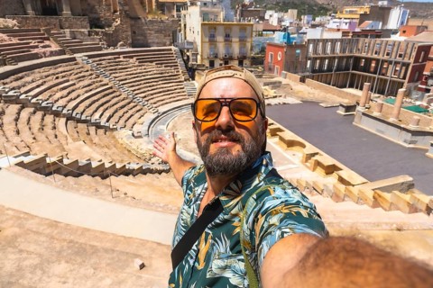Tourist man enjoying his vacation, taking a selfie while pointing to the ancient roman theatre in
