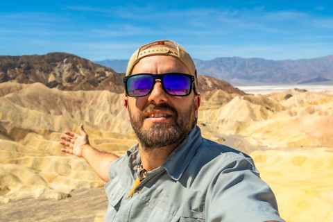 Man smiling at camera, making a welcoming gesture with his arm, showing the scenic desert landscape