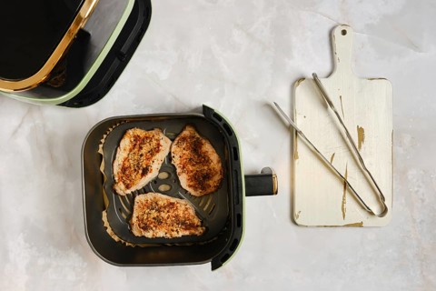 Pork chops grilled in air fryer. Kitchen tools like tongs and a cutting board are on side. The