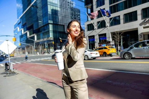 Young business woman smiling, holding a reusable coffee cup, and walking along a sunny city street