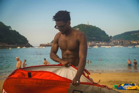 Young shirtless man on a sunny beach opening a vibrant red surf bag, preparing or packing up his