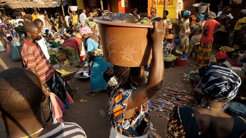 Woman wearing traditional clothes carrying a bucket on her head, lively market with lots of people