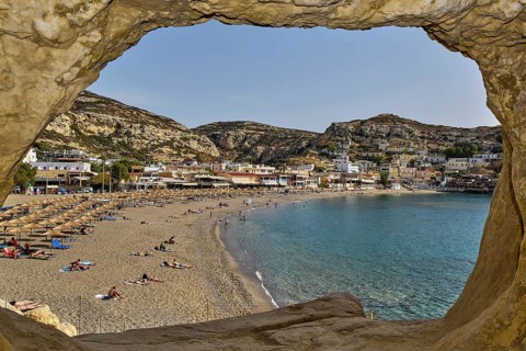 View of a busy beach with umbrellas and a village in the background, Matala, Neolithic caves,