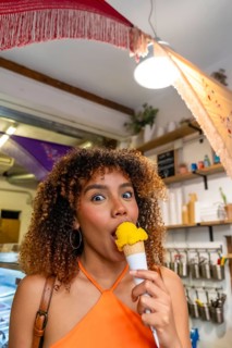 Young woman with curly hair happily eating a scoop of yellow ice cream from a waffle cone,
