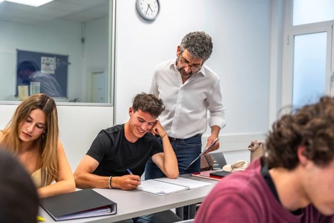 Teacher standing by a male student's desk, assisting him with his work, while other students are