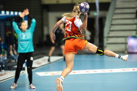 A female handball player in an orange jersey jumps to throw the ball towards the goal defended by a