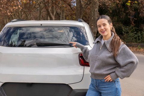 Smiling young woman pointing at rear windshield wiper area of white car, outdoor rural road, car