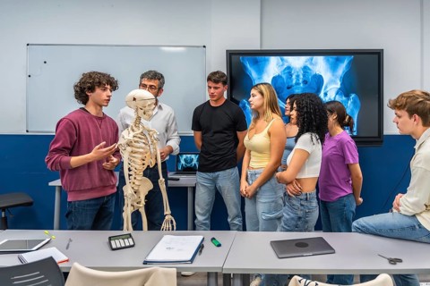 Group of high school students and their teacher analyzing a human skeleton model during an anatomy
