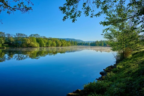 A wide view of a river with surrounding trees and clear skies, the calm water reflecting the sky,