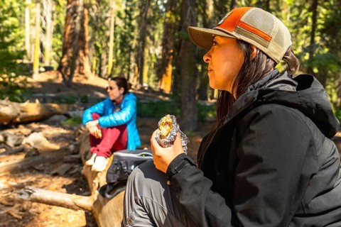 Woman in foreground eating a snack wrapped in foil, sitting outdoors in a forest clearing while a