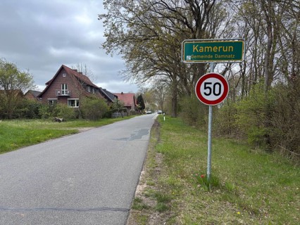 A rural road with speed limit and town sign, surrounded by houses and trees, Cameroon, Wendland,