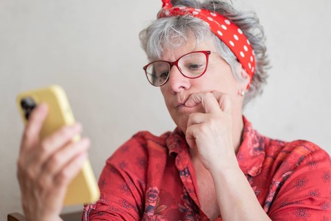 A senior woman wearing glasses and a red headband is holding and looking at a smartphone,