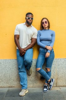 Cool modern multi-ethnic couple in denim clothes and fashion sunglasses leaning on urban wall