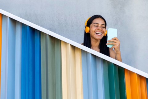 A joyful woman with headphones smiles while using her smartphone against a backdrop of vibrant