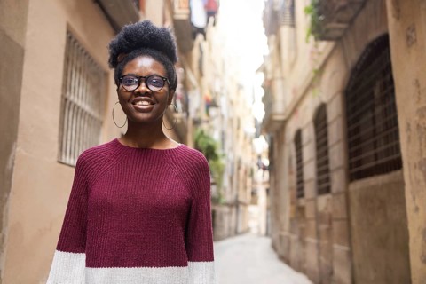 A cheerful woman wearing a burgundy sweater and glasses smiles confidently while standing in a