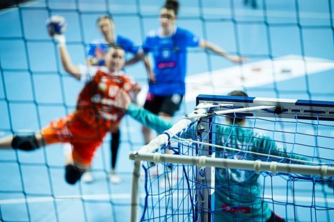 View through the handball net showing a blurred player in an orange jersey jumping to score a goal