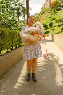 Smiling woman holding her small white dog while standing on a paved path in a park, surrounded by